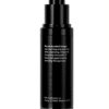 A black cosmetic spray bottle with a pump dispenser stands against a white background, reflecting on the surface below, with usage instructions printed on it.