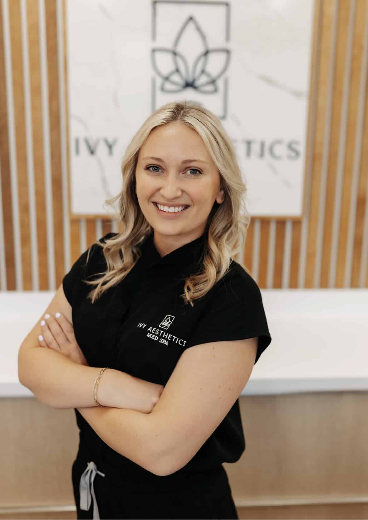 A smiling person in a black top stands with arms folded in front of a logo sign for "IVY AESTHETICS MED SPA," suggesting a professional setting.