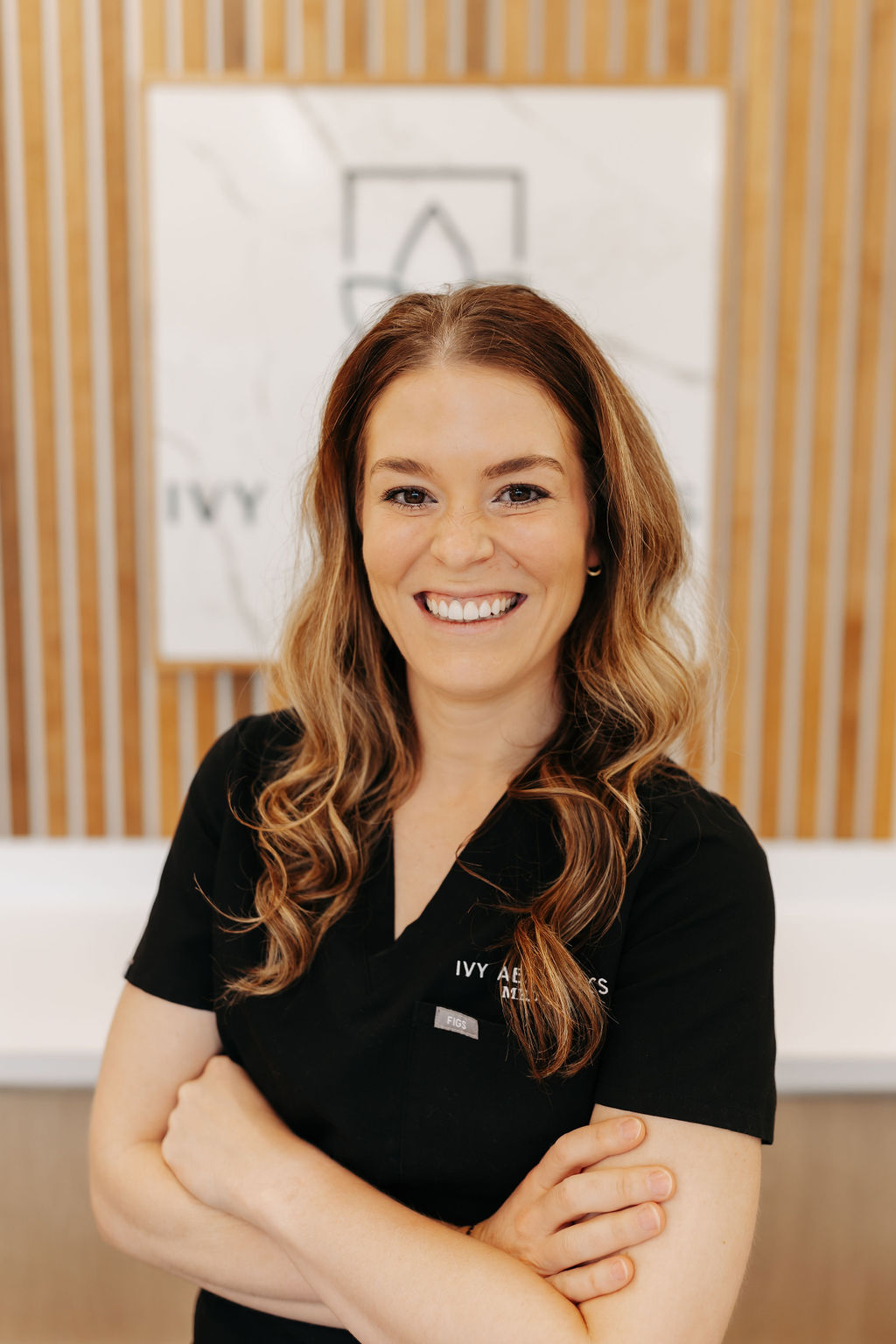 A smiling person in black attire stands in front of a wooden panel background, with a sign reading "IVY AESTHETICS" partially visible.