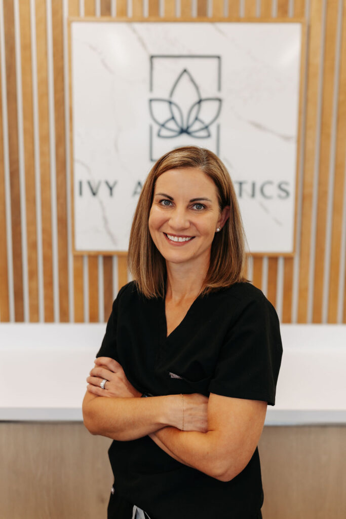 A smiling person in black attire stands with arms crossed, in front of a sign reading "IVY AESTHETICS" on a wooden background.