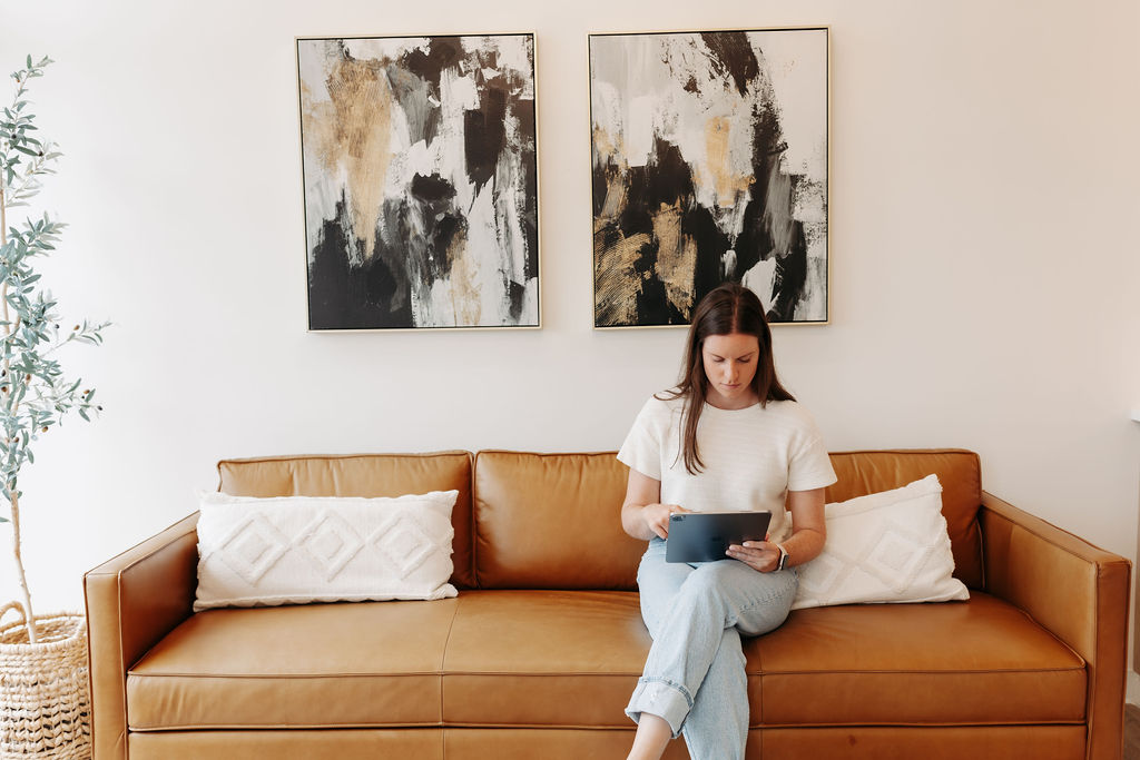 A person sits on a brown couch, holding a tablet. Abstract art hangs above, with a potted plant nearby in a modern setting.