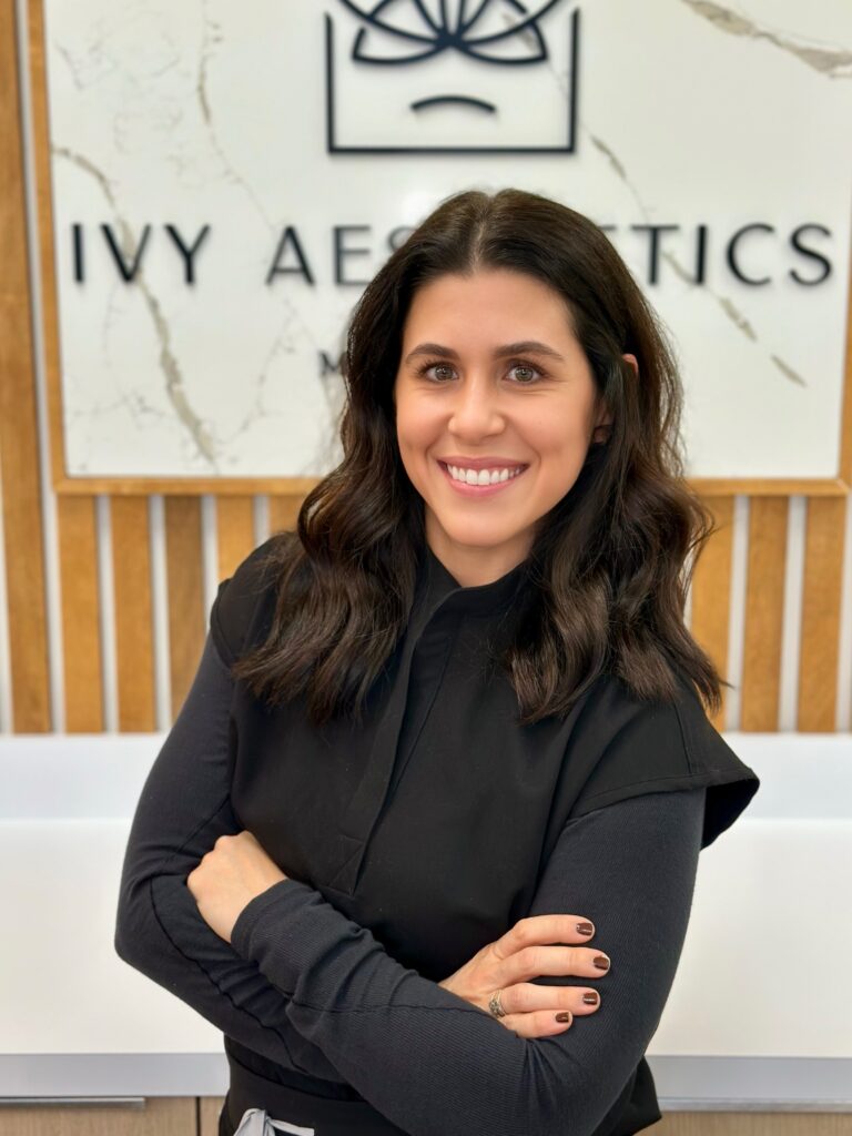 A person with dark hair smiles confidently, arms crossed, in front of an "Ivy Aesthetics" sign on a wooden and marble background.