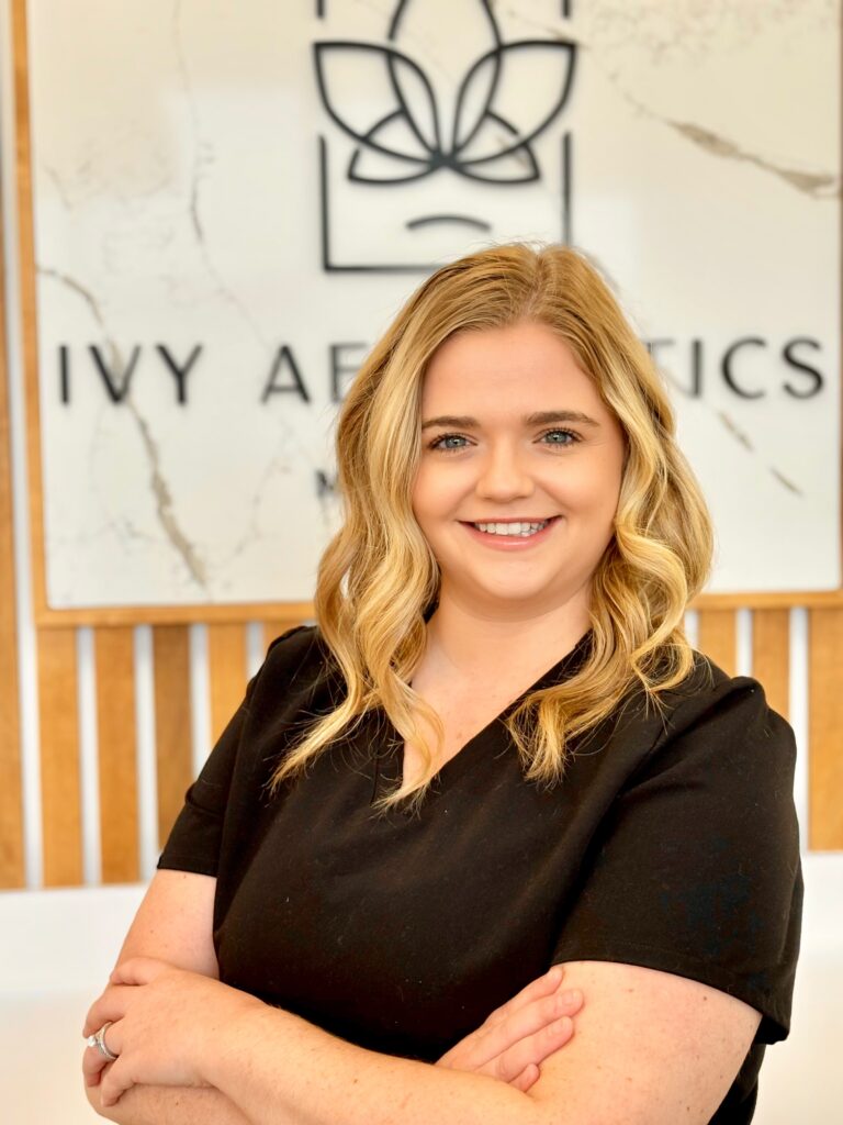 A smiling person with blonde hair stands in front of a sign reading "Ivy Aesthetics," wearing a black top, arms crossed confidently.
