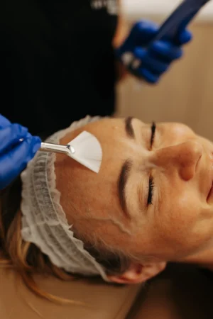A person receiving a facial treatment, smiling with closed eyes. Blue-gloved hand applies skincare with a brush to the forehead.
