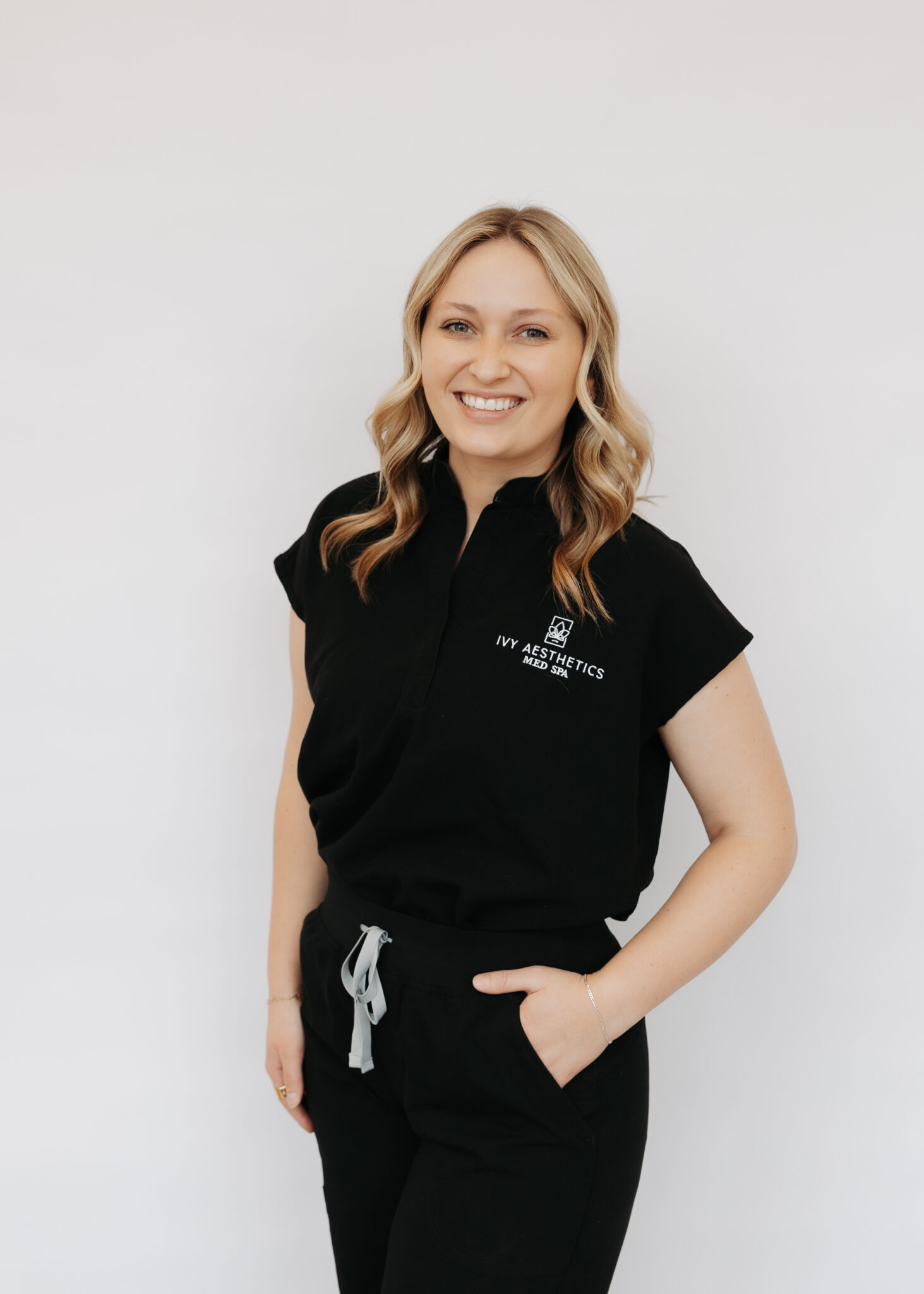 A person with blonde hair smiles, wearing a black outfit with "Ivy Aesthetics Med Spa" logo against a plain white background.