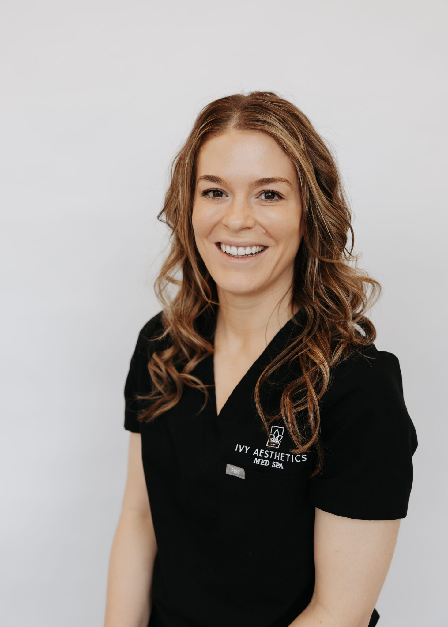 A person with wavy hair smiles, wearing a black uniform with "Ivy Aesthetics Med Spa" logo on a plain white background.