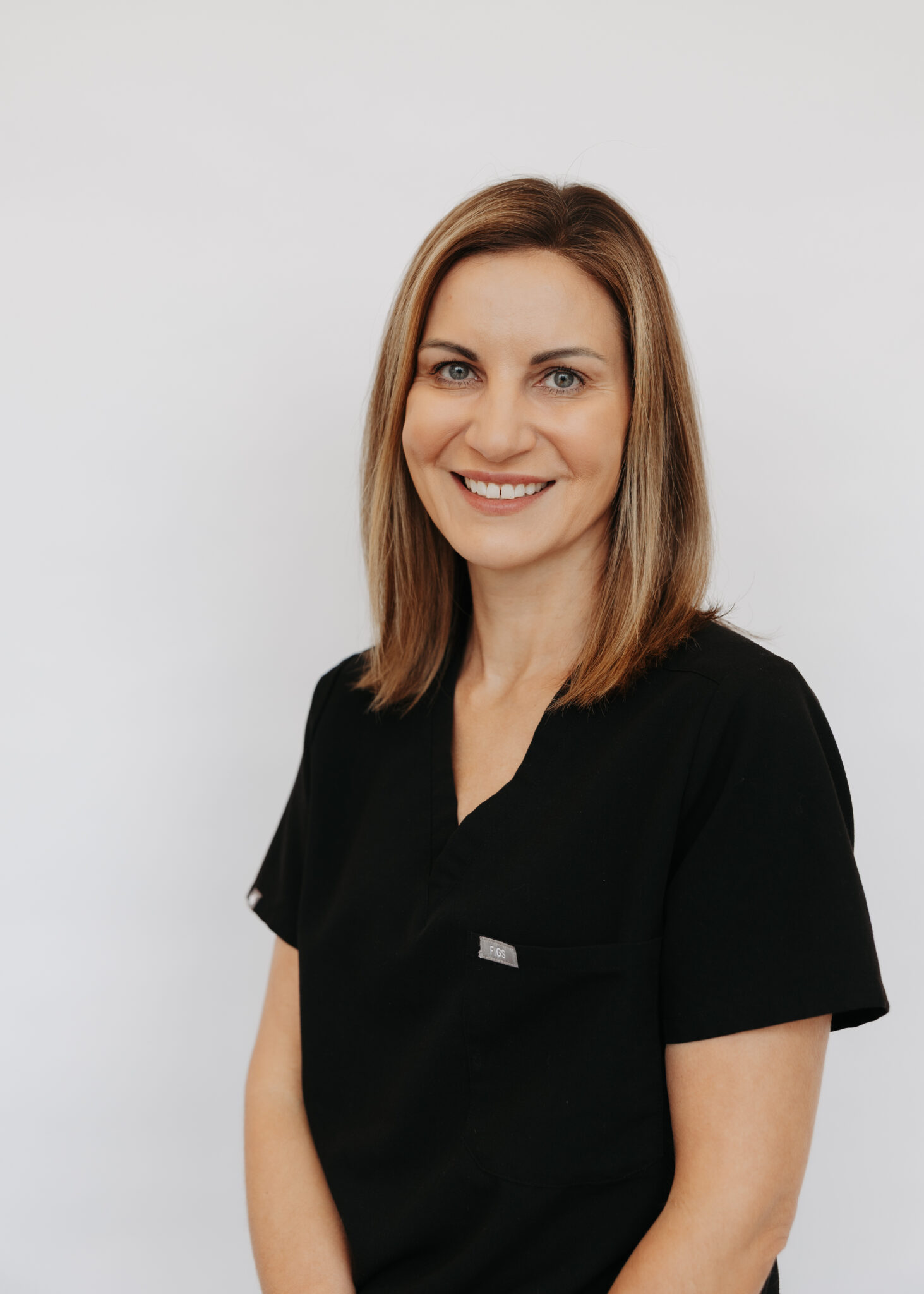 A person wearing black scrubs smiles against a plain white background, conveying a professional and approachable demeanor in this portrait photograph.