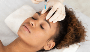 A person lies relaxed as another administers a forehead injection with blue liquid, wearing white gloves, indicating a cosmetic or medical procedure.