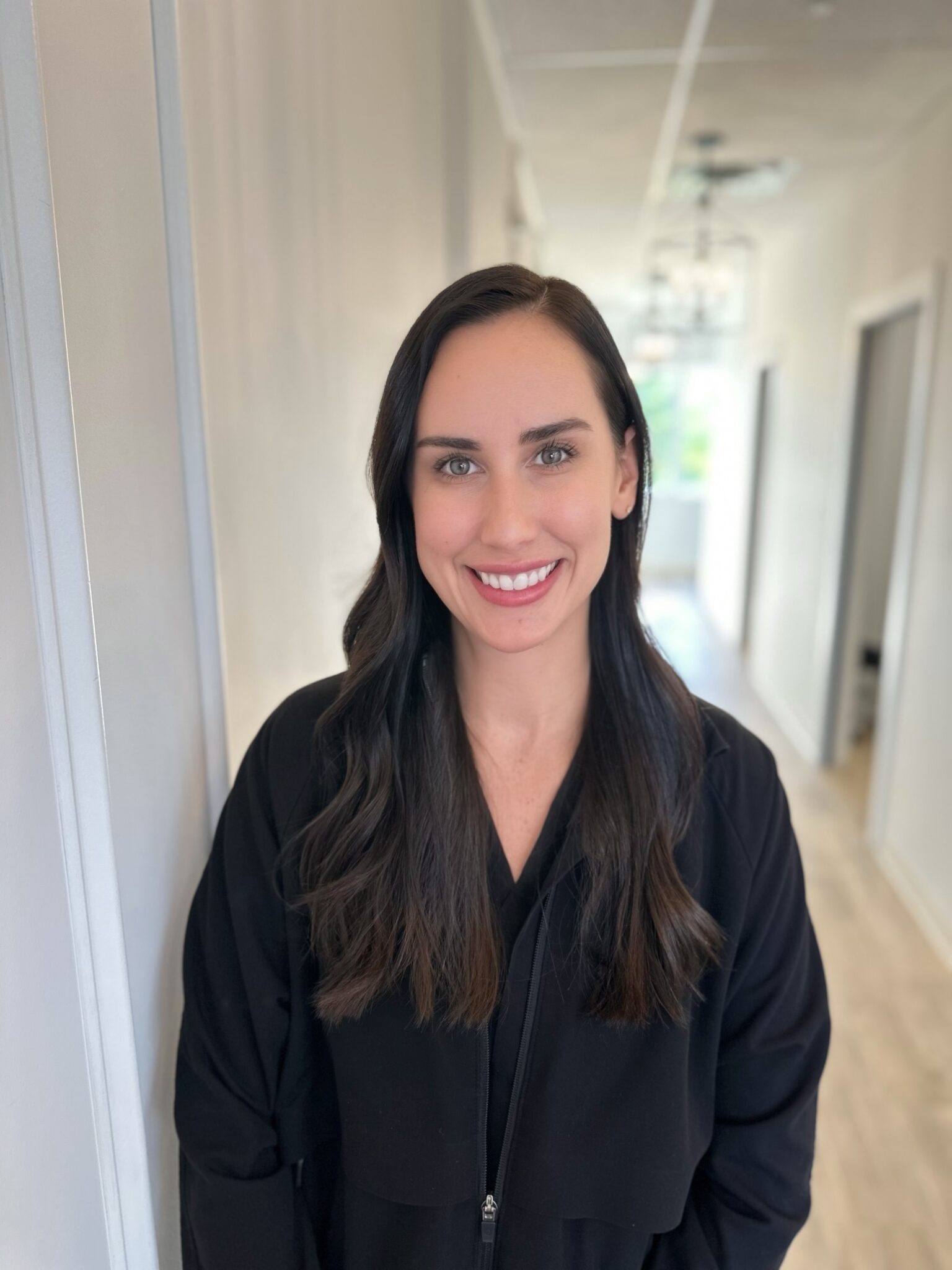 A person with long dark hair in a black outfit smiles in a brightly lit, modern hallway with a wooden floor.