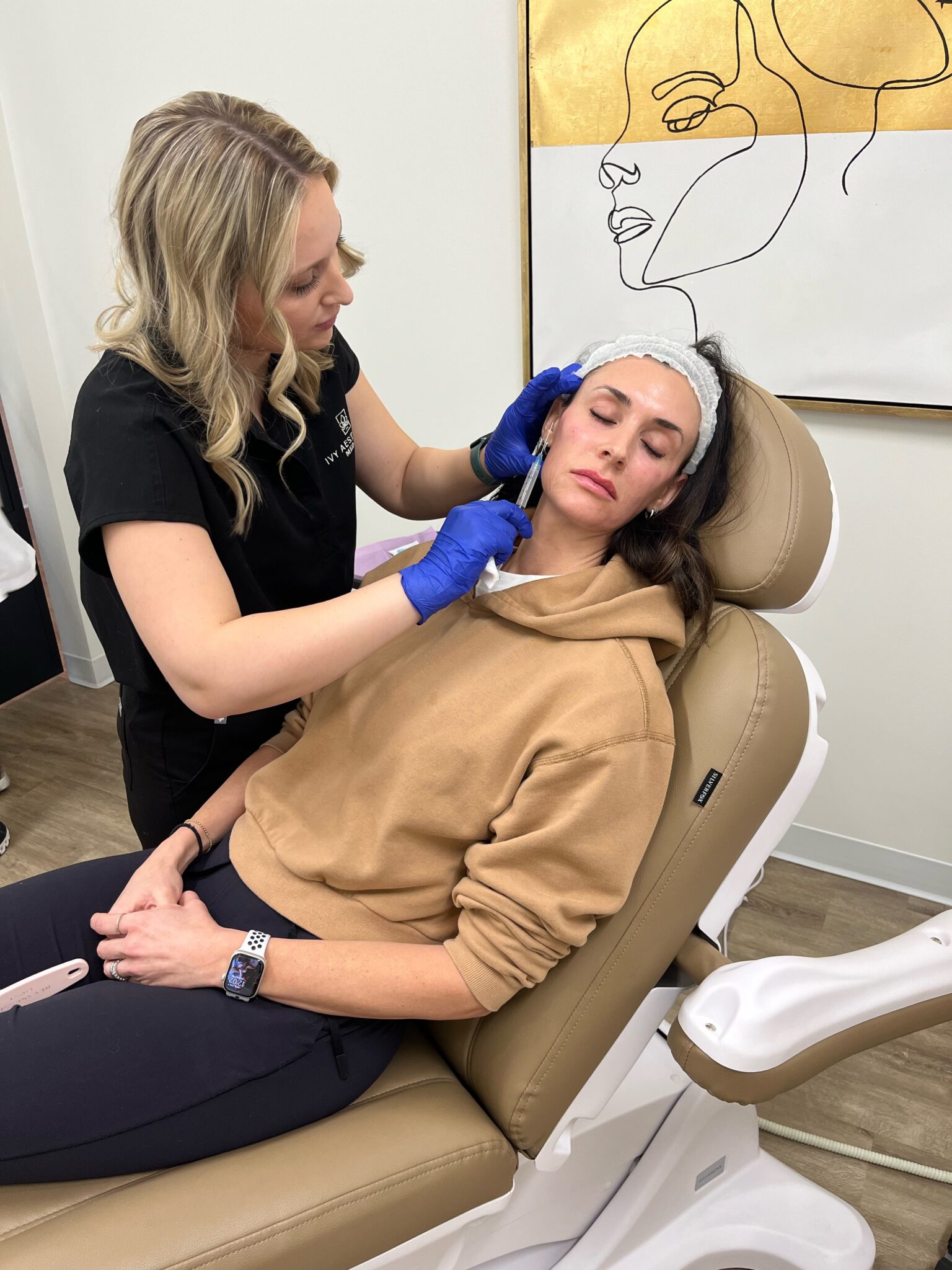 A person in a medical setting receives a facial injection from another person, with a line-art portrait on the wall.