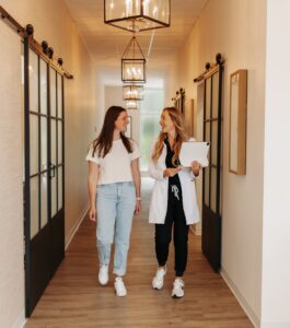 Two people walking and talking in a well-lit hallway, one holding a tablet. Modern decor with hanging lights and sliding glass doors.