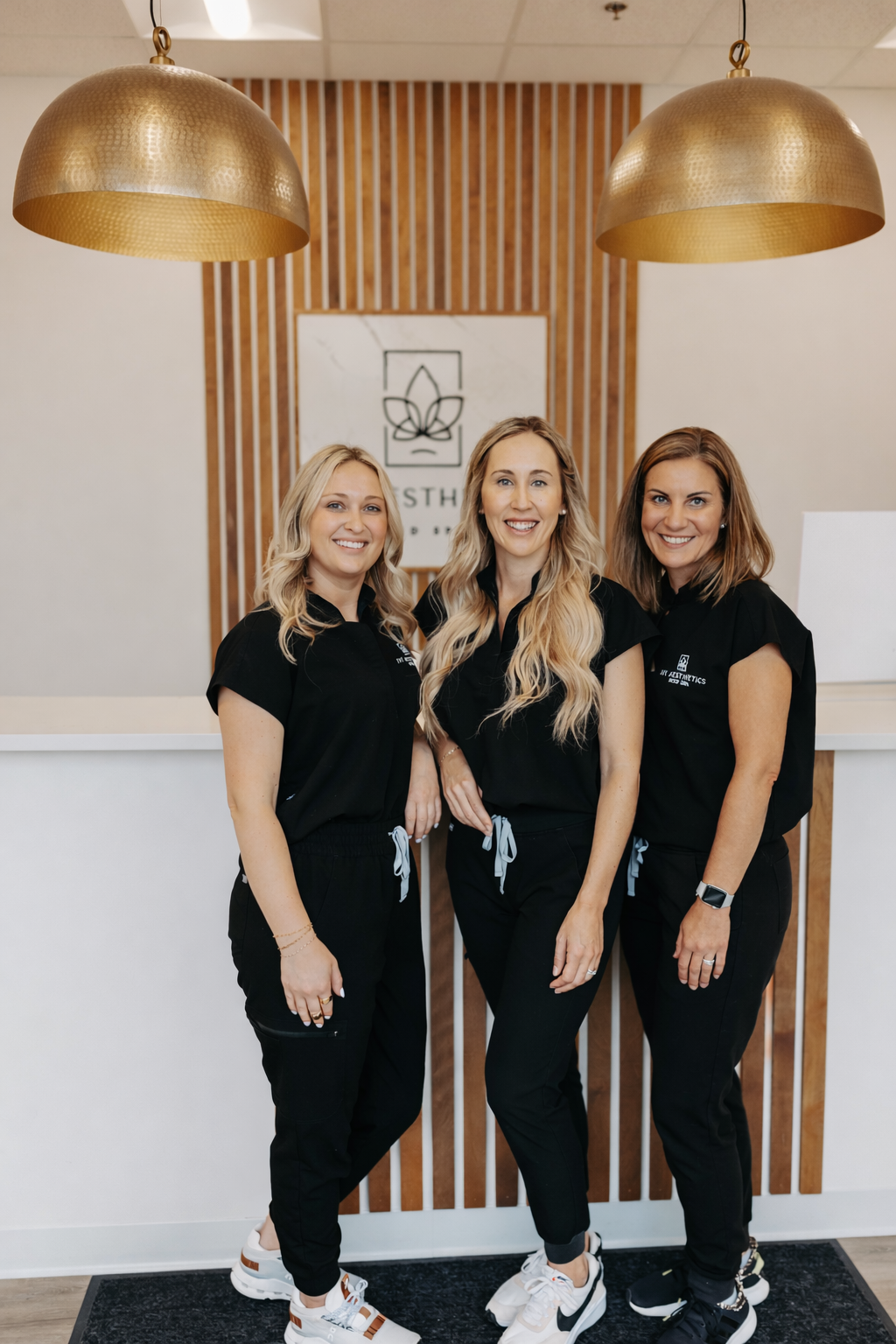 Three people in black outfits smile in a modern office with large hanging lights and a sign in the background.