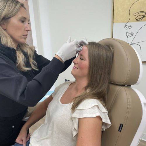 A person is performing a cosmetic procedure on another person's forehead in a clinical setting with modern decor, including a framed abstract artwork.