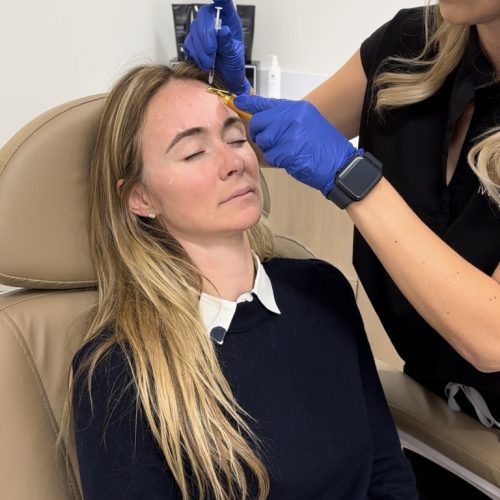 A person in a black sweater sits in a chair while another person wearing blue gloves applies a treatment between the seated person's eyebrows.