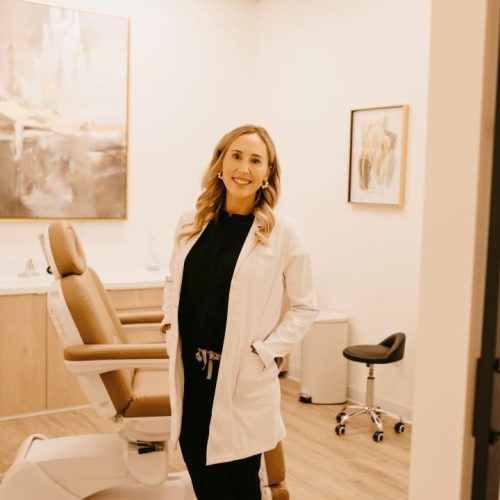 A smiling person in a white lab coat stands confidently in a dental office with a patient chair and elegant interior decoration in the background.