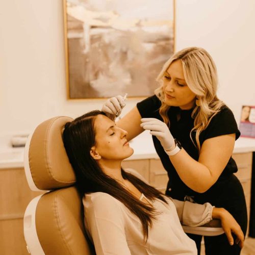One person is receiving a beauty treatment from another, who is wearing gloves, in a salon-like setting with a neutral color palette.
