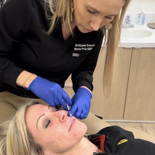A person in a black uniform marked 'Nurse Practitioner' examines another reclining person's face, wearing gloves, in a clinical setting with medical equipment.