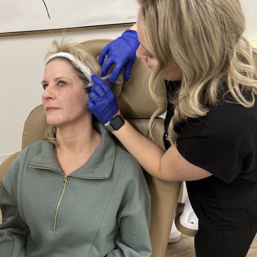 A person wearing gloves inspects another seated person's head, with markings on their face, possibly for a cosmetic procedure, in a clinic-like setting.