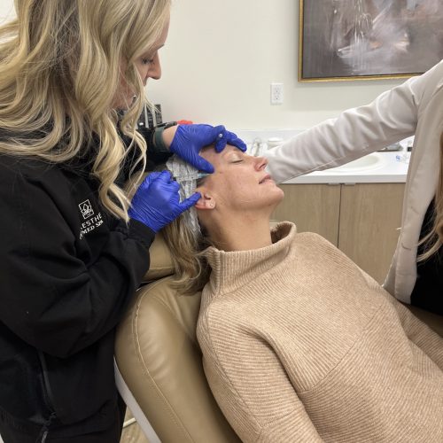 A seated person receives a facial treatment from a standing person wearing gloves in a clinical setting. A piece of artwork hangs on the wall behind.