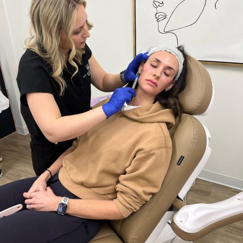 A person is performing a facial treatment on another seated person in a clinical setting, surrounded by medical or aesthetic equipment and artwork.